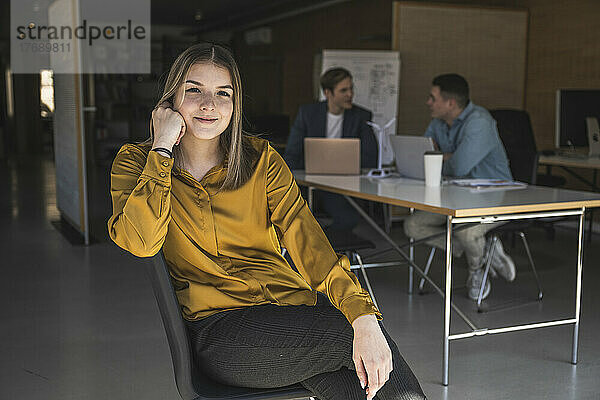 Selbstbewusste Geschäftsfrau sitzt auf einem Stuhl im Büro mit Kollegen im Hintergrund