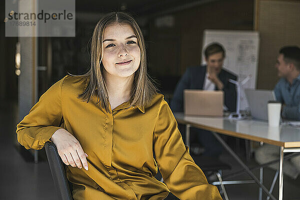 Selbstbewusste Geschäftsfrau sitzt auf einem Stuhl im Büro mit Kollegen im Hintergrund