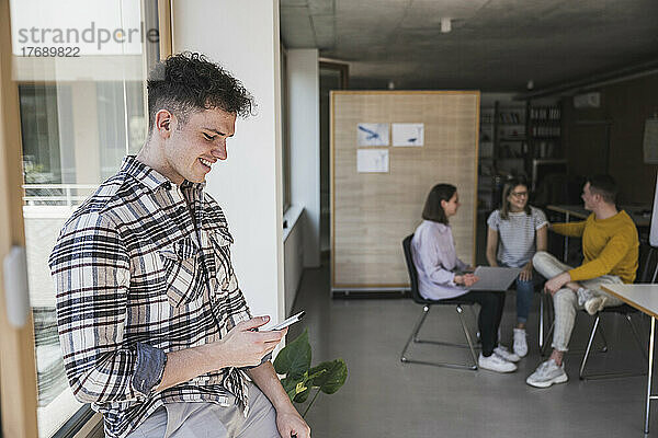 Lächelnder junger Geschäftsmann nutzt Smartphone im Büro mit Kollegen im Hintergrund