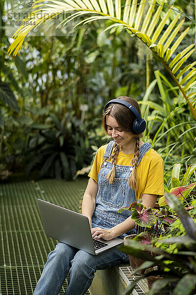Glückliche Frau benutzt Laptop und hört Musik über kabellose Kopfhörer im Garten