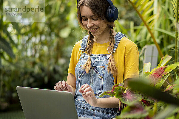 Glückliche junge Frau mit kabellosen Kopfhörern sitzt mit Laptop im Garten