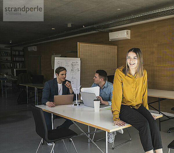 Glückliche Geschäftsfrau sitzt am Tisch im Büro mit Kollegen im Hintergrund