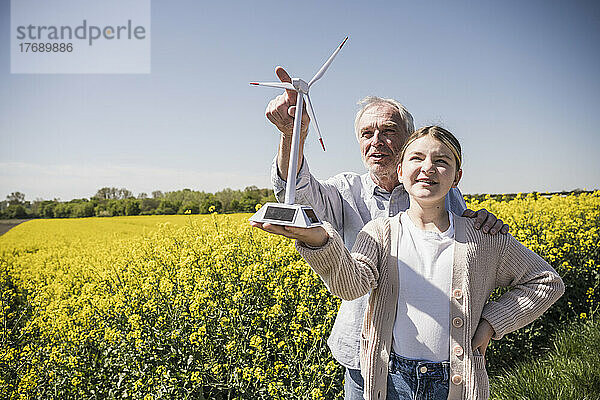 Mädchen hält Modell einer Windkraftanlage in der Hand  während Großvater auf landwirtschaftliches Feld gestikuliert