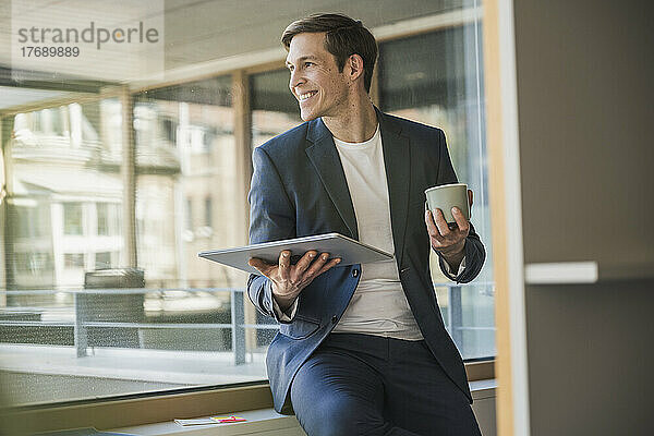 Selbstbewusster Geschäftsmann hält Tablet und Kaffeetasse am Fenster im Büro
