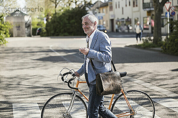 Lächelnder Geschäftsmann mit Fahrrad überquert Straße an sonnigem Tag