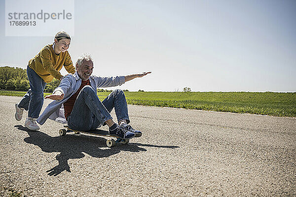 Verspielte Enkelin schiebt den glücklichen Großvater  der auf dem Skateboard sitzt