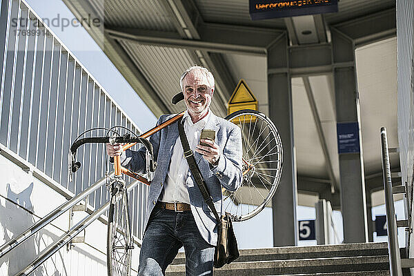 Lächelnder Geschäftsmann  der ein Fahrrad auf der Schulter trägt und sein Mobiltelefon in der Hand hält und sich von der Treppe hinunterbewegt
