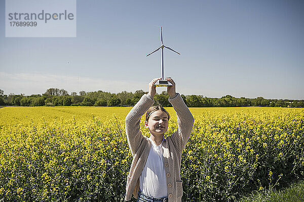 Lächelndes Mädchen mit geschlossenen Augen hält ein Modell einer Windkraftanlage  die auf dem Feld steht