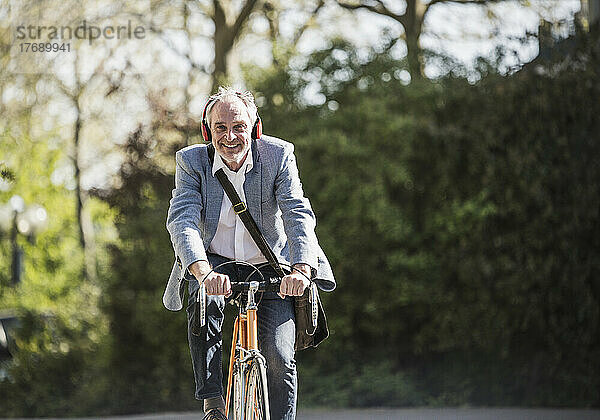 Lächelnder Geschäftsmann  der an einem sonnigen Tag Musik über drahtlose Kopfhörer beim Radfahren hört
