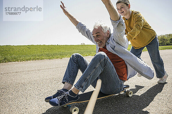 Verspieltes Mädchen schiebt fröhlichen Großvater  der mit erhobenen Armen auf dem Skateboard sitzt