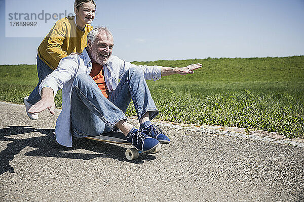 Glückliches Mädchen schubst Großvater  der mit ausgestreckten Armen auf dem Skateboard sitzt