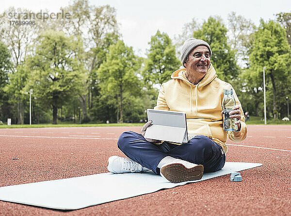 Lächelnder älterer Mann mit Tablet-PC und Wasserflasche sitzt auf einer Trainingsmatte