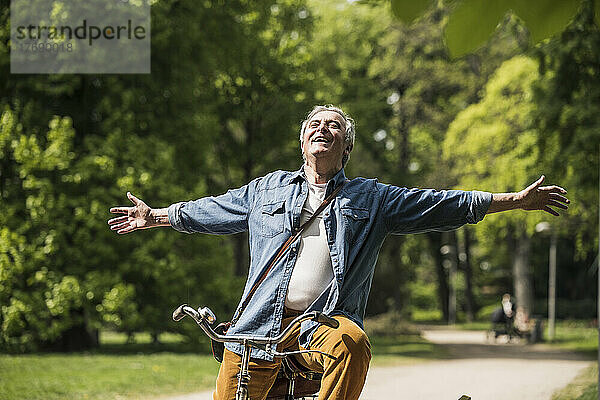 Sorgloser älterer Mann mit ausgestreckten Armen  der im Park Fahrrad fährt