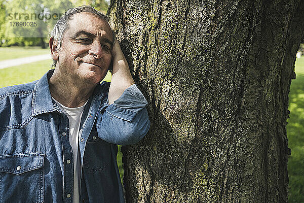 Lächelnder älterer Mann lehnt im Park an Baumstamm