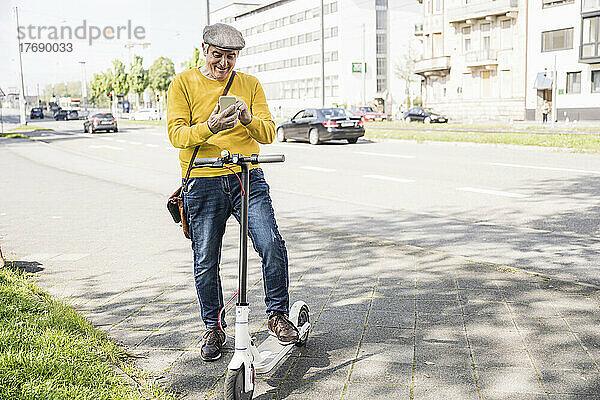 Glücklicher älterer Mann  der sein Mobiltelefon benutzt und mit einem Elektroroller steht