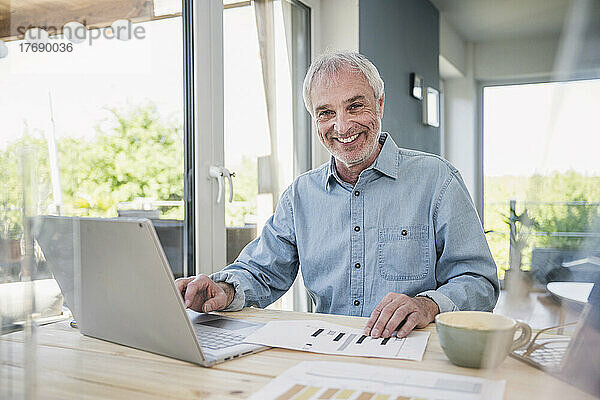 Glücklicher älterer Geschäftsmann mit Laptop und Grafik  der am Tisch sitzt