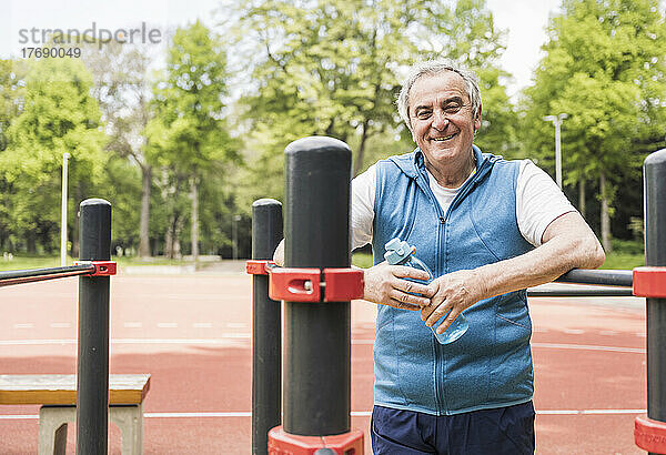 Lächelnder älterer Mann hält eine Wasserflasche in der Hand und steht inmitten einer Gymnastikbar