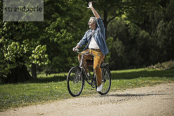 Lächelnder älterer Mann  der an einem sonnigen Tag Fahrrad fährt und im Park mit der Hand winkt