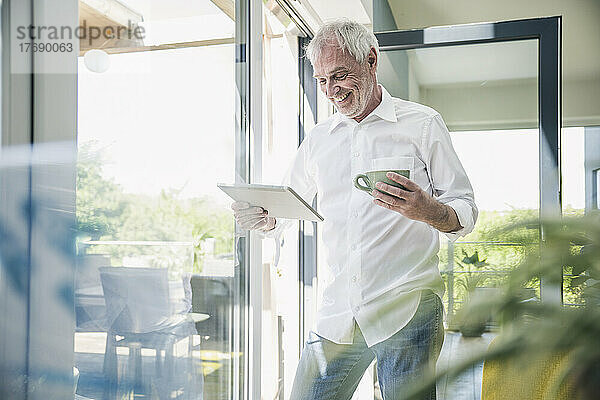 Glücklicher älterer Mann mit Tablet-PC und Kaffeetasse am Glasfenster