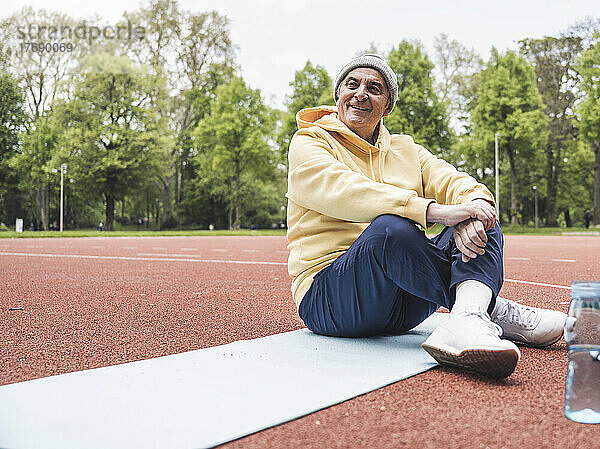 Glücklicher älterer Mann mit gefalteten Händen  der mit gekreuzten Beinen auf einer Trainingsmatte im Park sitzt