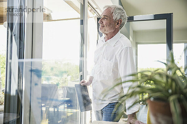 Smiling senior man with tablet PC leaning on table day dreaming at home