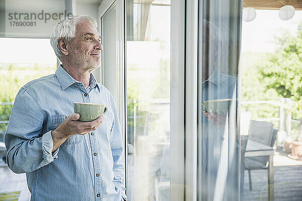 Glücklicher älterer Mann mit Kaffeetasse und Blick aus dem Fenster