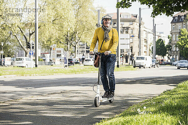 Lächelnder älterer Mann mit kabellosen Kopfhörern  der auf einem elektrischen Tretroller fährt