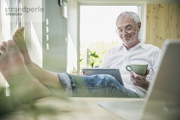 Lächelnder älterer Mann mit Kaffeetasse und Tablet-PC  der zu Hause sitzt