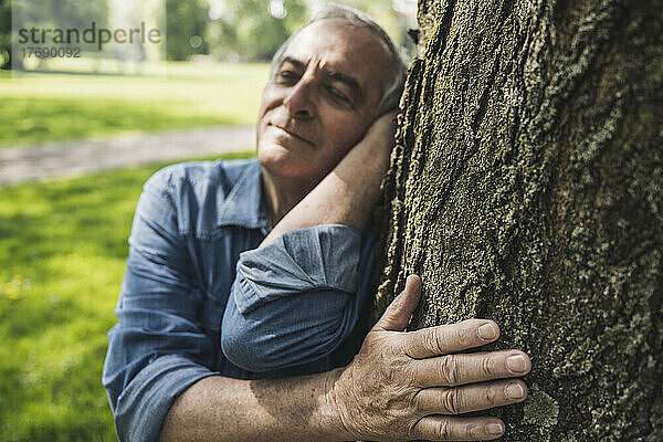 Rentner älterer Mann mit geschlossenen Augen lehnt an Baumstamm im Park