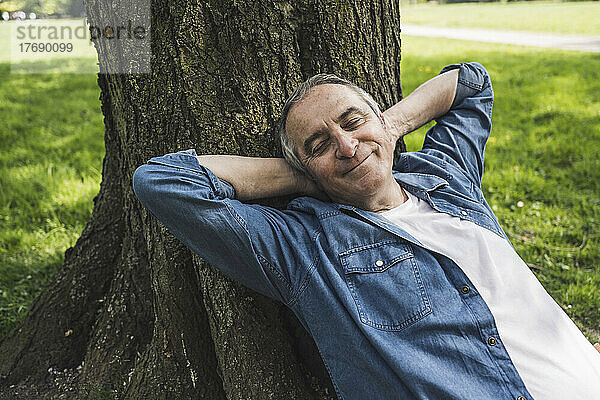 Lächelnder älterer Mann im Ruhestand mit geschlossenen Augen  der sich im Park an einen Baumstamm lehnt