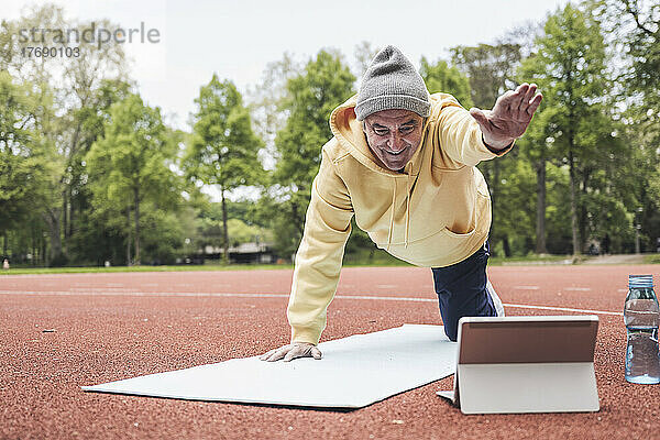 Glücklicher älterer Mann  der im Park eine E-Learning-Yoga-Übung aus einem Tutorial auf einem Tablet-PC durchführt