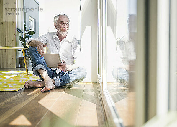 Lächelnder älterer Mann mit Tablet-PC sitzt zu Hause auf dem Boden