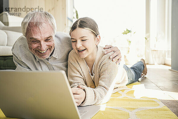 Glücklicher älterer Mann mit Enkelin  der zu Hause auf dem Teppich im Wohnzimmer liegt und einen Laptop benutzt