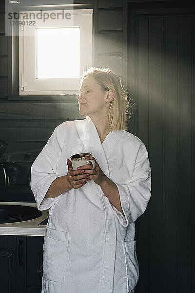 Frau im Bademantel hält Tasse in der Hand und genießt zu Hause das Sonnenlicht
