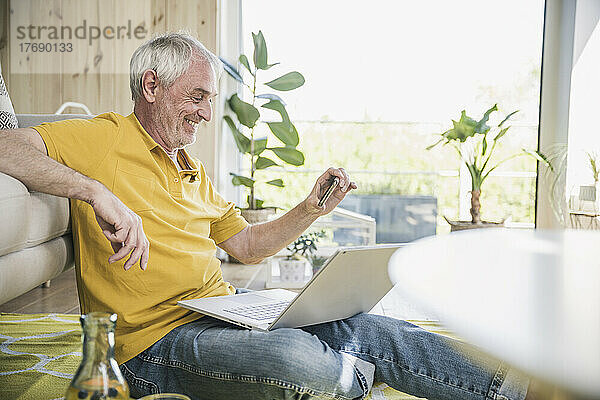 Smiling senior man using smart phone sitting with laptop at home