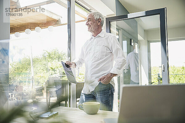 Lächelnder älterer Mann mit Tablet-PC vor Glasfenster zu Hause