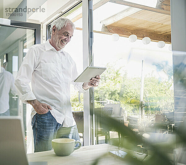 Glücklicher älterer Mann mit Tablet-PC  der zu Hause am Glasfenster steht