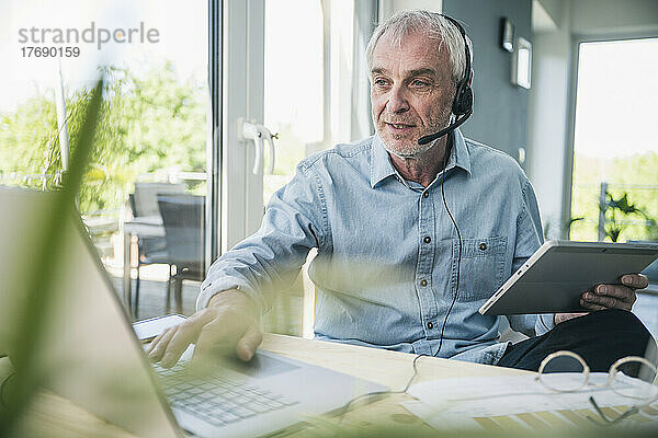 Älterer Geschäftsmann trägt Headset und arbeitet zu Hause am Laptop