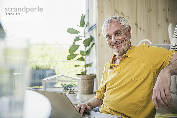 Lächelnder älterer Mann mit Laptop sitzt zu Hause im Wohnzimmer