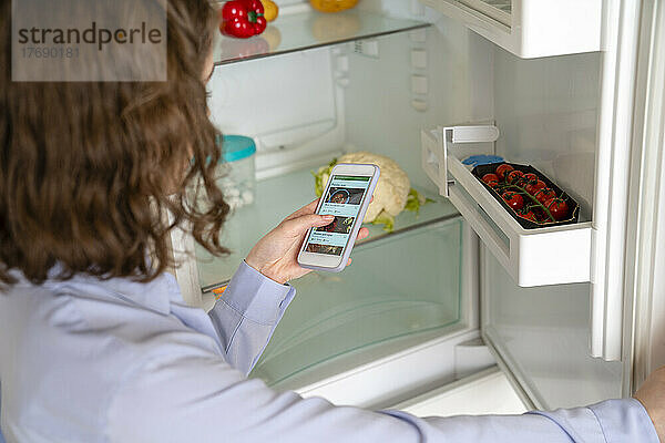 Frau kauft online Lebensmittel per Smartphone am Kühlschrank ein