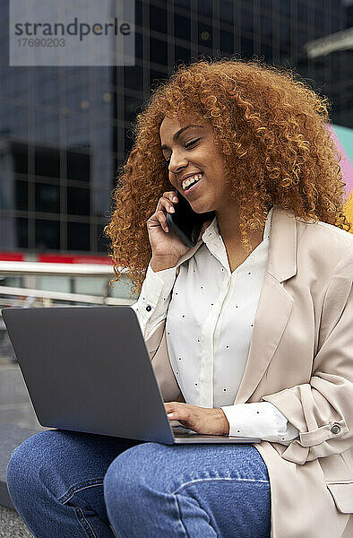 Lächelnde Geschäftsfrau mit Laptop  die auf dem Smartphone spricht
