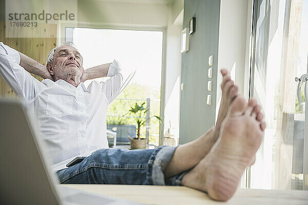 Entspannter älterer Mann sitzt mit den Händen hinter dem Kopf und gekreuzten Beinen am Tisch
