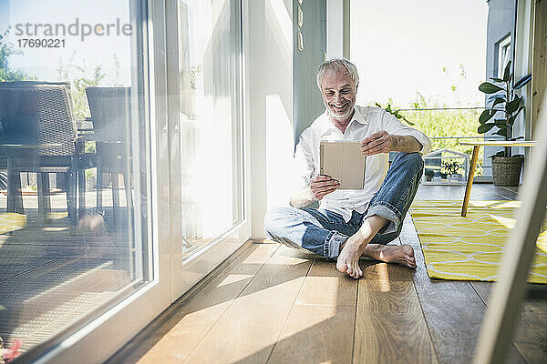 Glücklicher älterer Mann mit Tablet-PC  der zu Hause auf dem Boden im Wohnzimmer sitzt