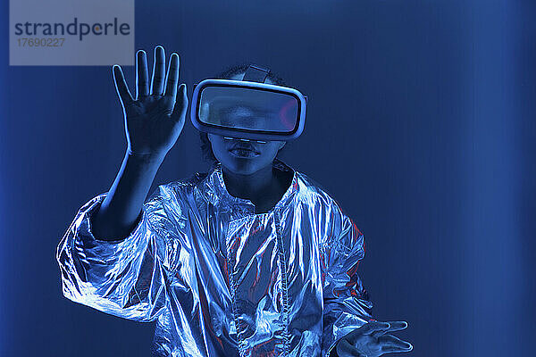 Junge Frau im Virtual-Reality-Simulator gestikuliert vor blauem Hintergrund