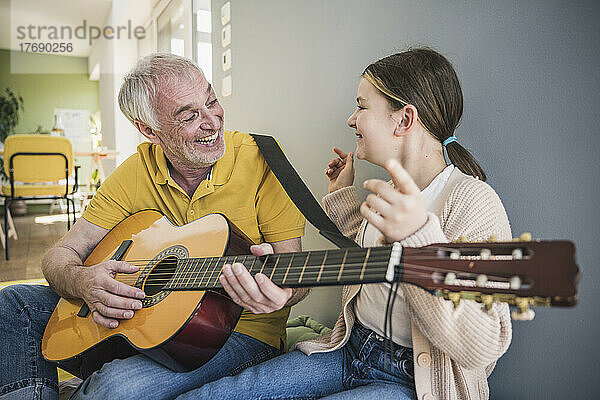Glücklicher älterer Mann  der zu Hause von seiner Enkelin Gitarre spielt