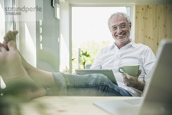Glücklicher älterer Mann mit Kaffeetasse und Tablet-PC  der zu Hause sitzt