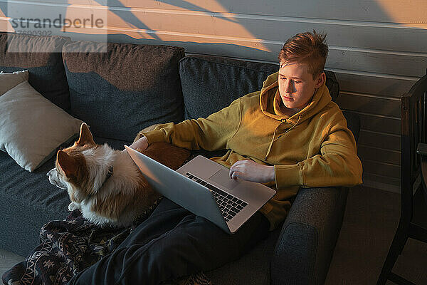 Junge benutzt Laptop und sitzt mit Hund zu Hause auf dem Sofa