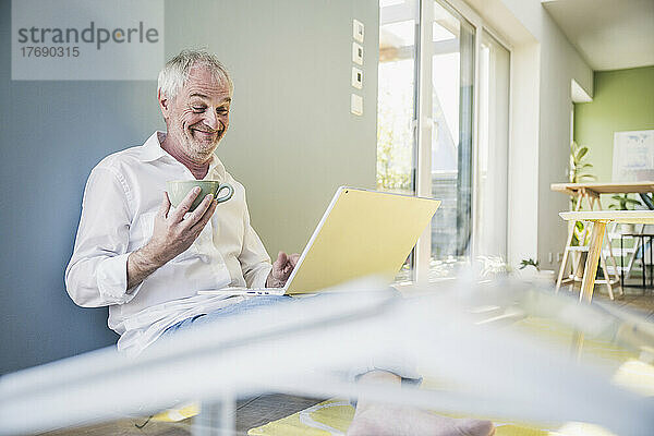 Glücklicher älterer Mann  der zu Hause eine Kaffeetasse mit einem Laptop in der Hand hält