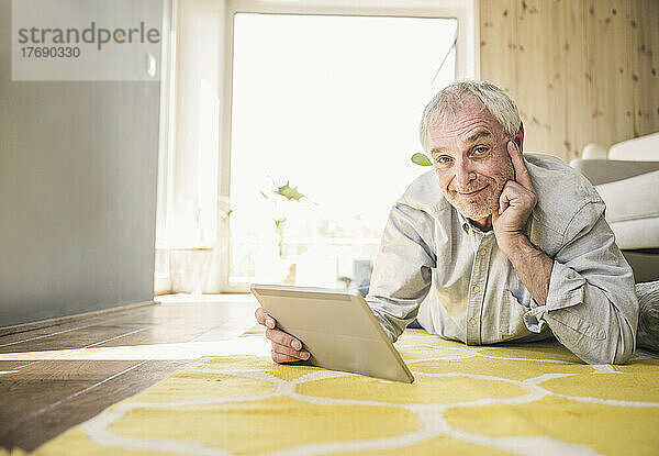 Lächelnder älterer Mann mit Tablet-PC  der zu Hause auf dem Teppich liegt