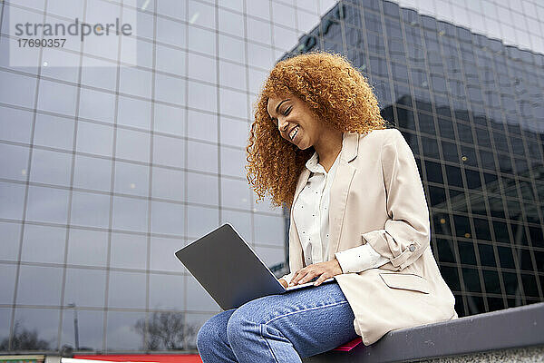 Lächelnde Geschäftsfrau  die vor dem Bürogebäude am Laptop arbeitet